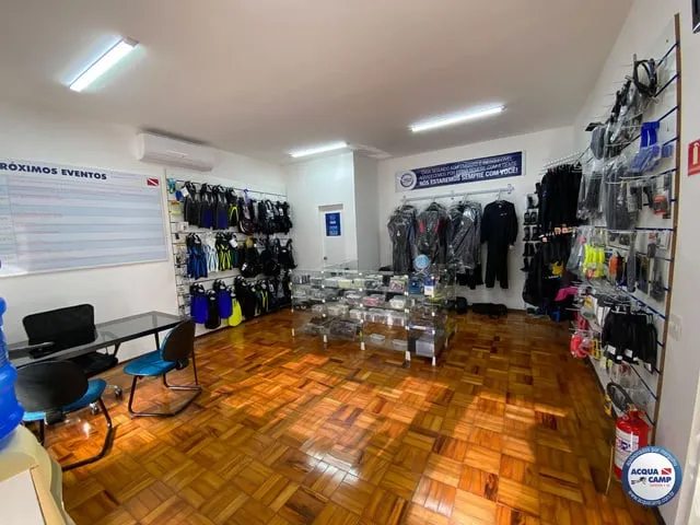 Interior da loja da Acquacamp: roupas de neoprene e equipamento nas paredes, piso de tacos e o quadro de próximos eventos