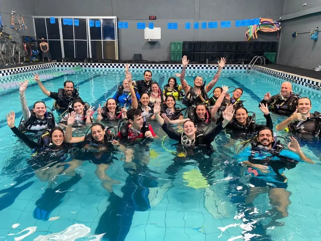 Cerca de vinte alunos de roupa de neoprene dentro da piscina aquecida da Acquacamp, acenando para a câmera