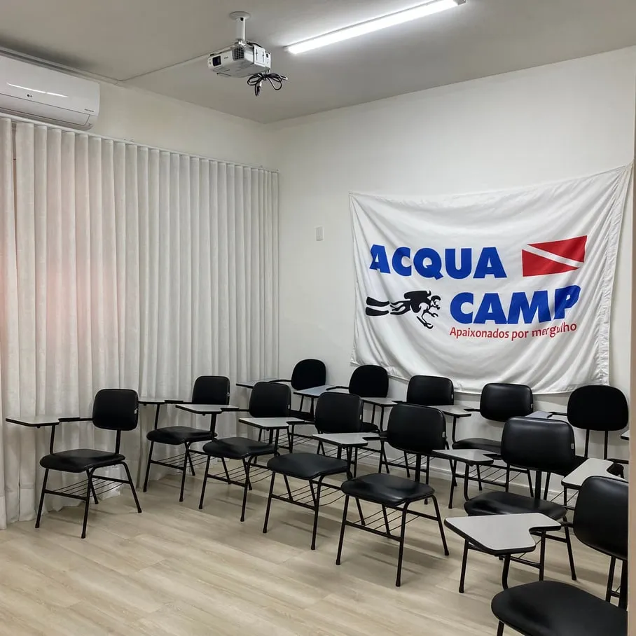 Sala de aula da Acquacamp com cadeiras universitárias, projetor no teto, ar condicionado e a bandeira da escola na parede